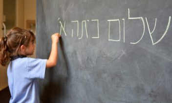Little girl (Age 5-6) writes Hello First Grade greetings in Hebrew (Shalom Kita Alef) on a chalkboard in Israeli primary school at the beginning of the school year. Education concept photo